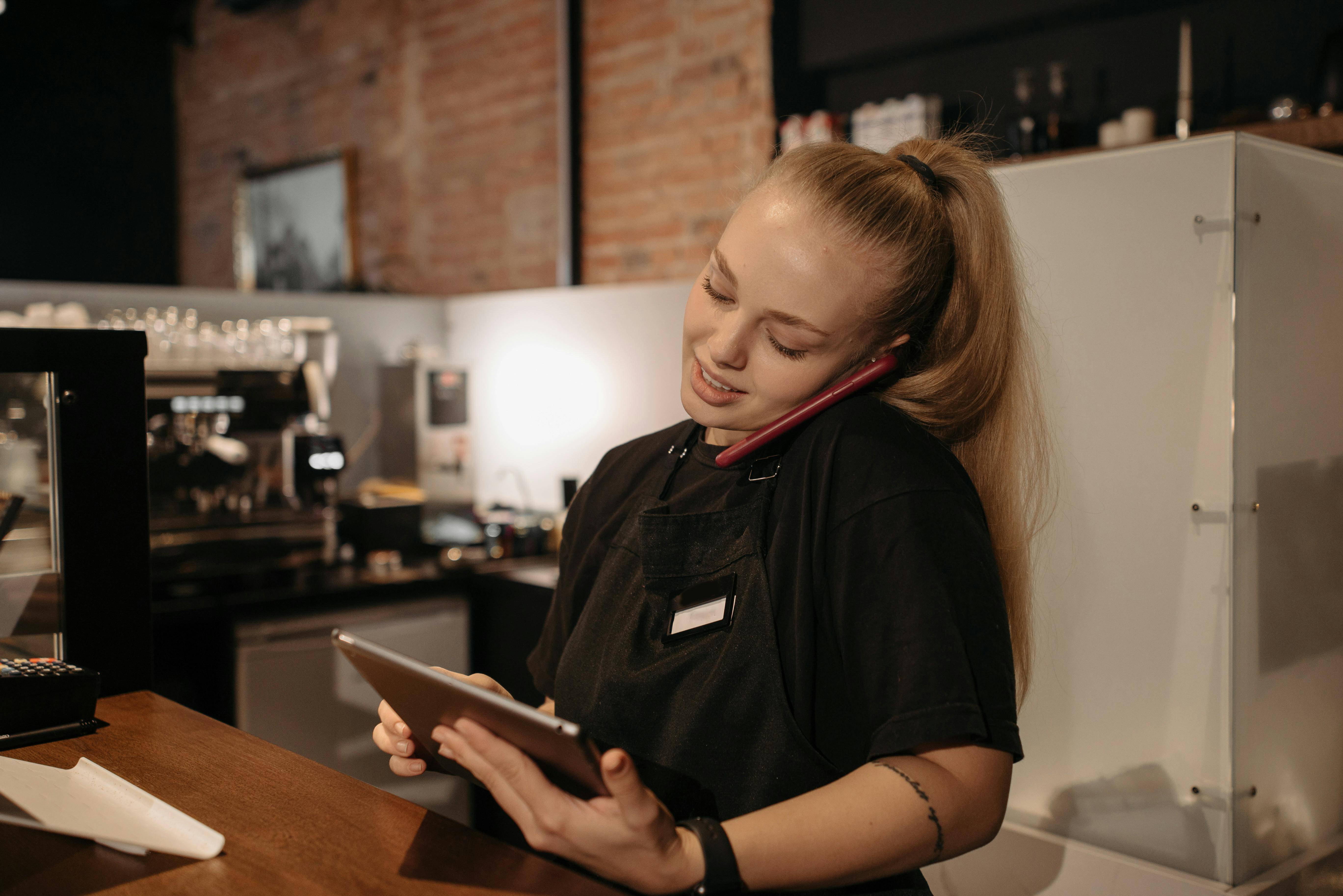 An employee behind the counter of a restaurant who is on the phone and working on a tablet at the same time