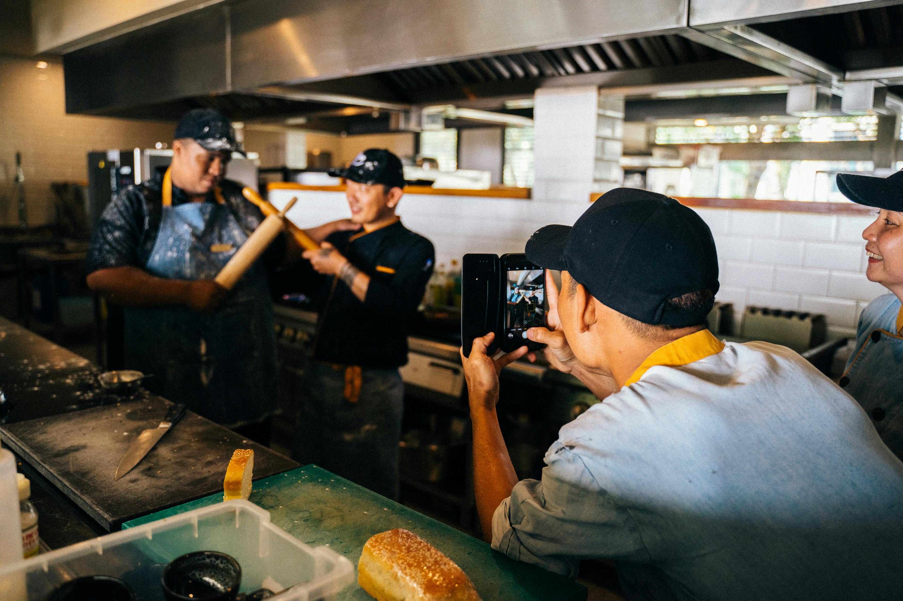 An employee takes a video of the kitchen team holding the rolling pins in the kitchen.